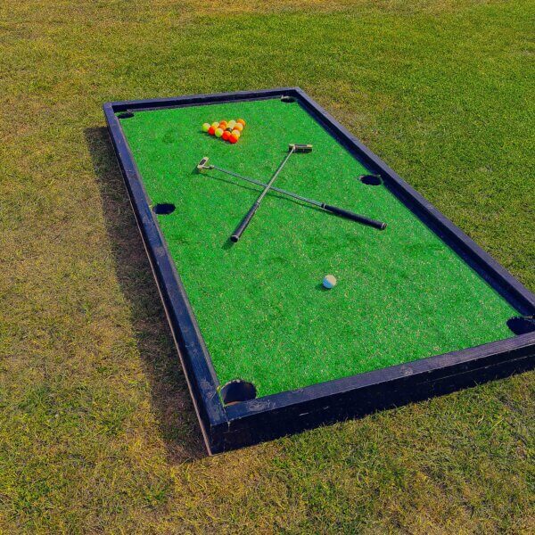 Large golf pool tables on the floor with red and yellow balls arranged in a triangle with a middle black ball. A white ball sits at the other end with 2 criss-crossed golf putters
