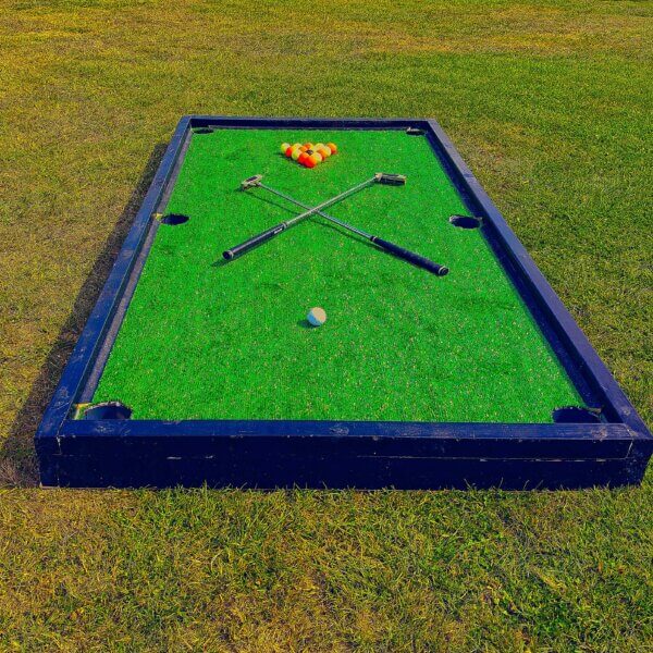 Large golf pool tables on the floor with red and yellow balls arranged in a triangle with a middle black ball. A white ball sits at the other end with 2 criss-crossed golf putters