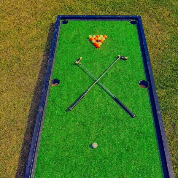 Large golf pool tables on the floor with red and yellow balls arranged in a triangle with a middle black ball. A white ball sits at the other end with 2 criss-crossed golf putters