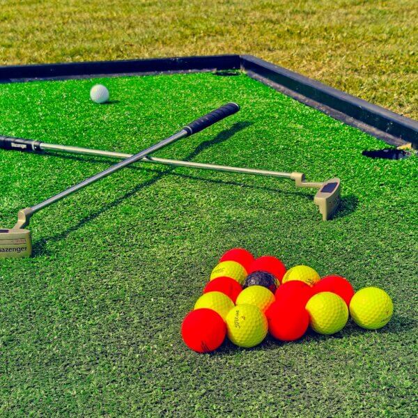 Large golf pool tables on the floor with red and yellow balls arranged in a triangle with a middle black ball. A white ball sits at the other end with 2 criss-crossed golf putters