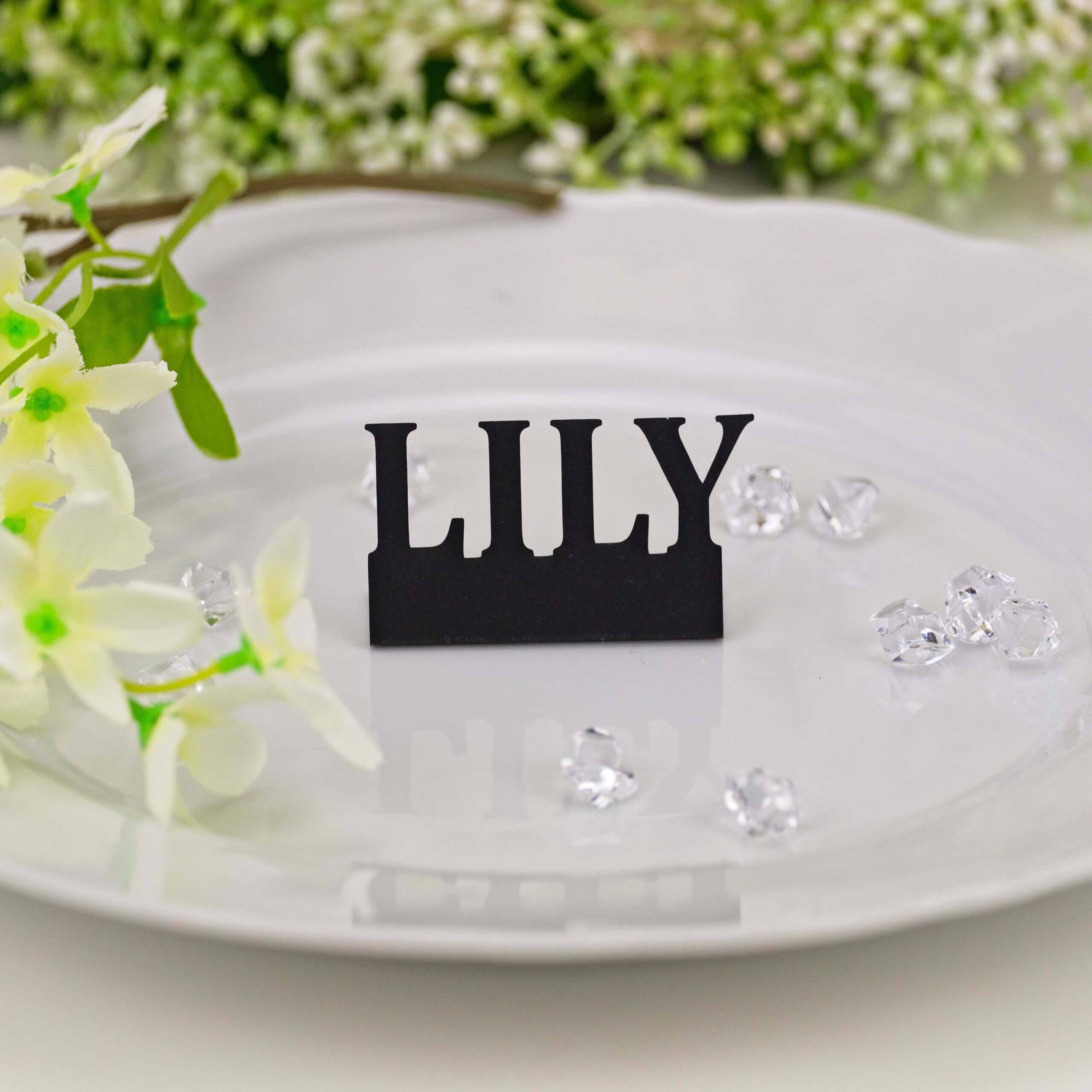 Black "LILY" standing name cut-out on white plate with flowers and crystal accents, close-up wedding table shot.