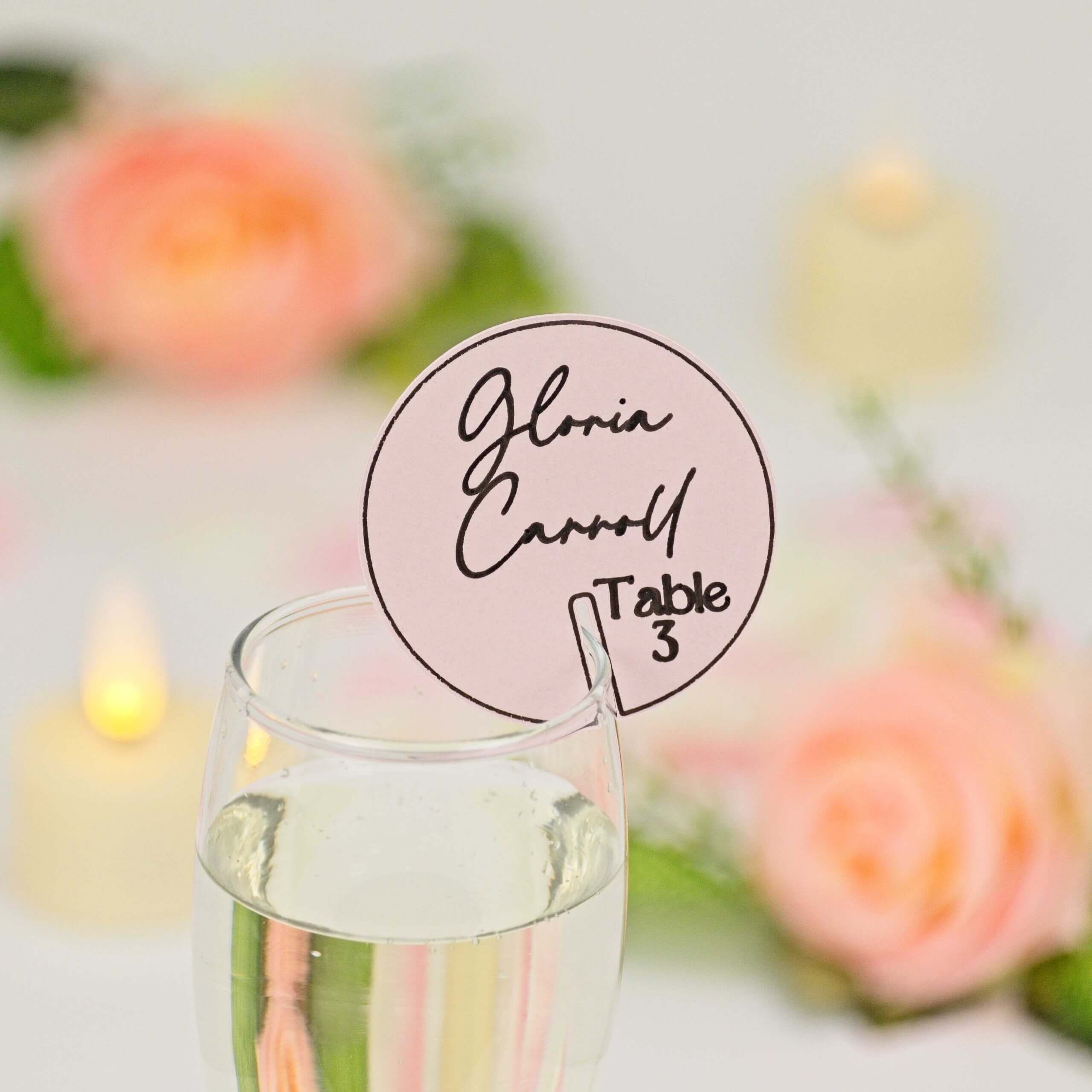 Round blush-pink drink tag with black script guest name and "Table 3", clipped to a champagne flute; soft-focus floral and candle background.