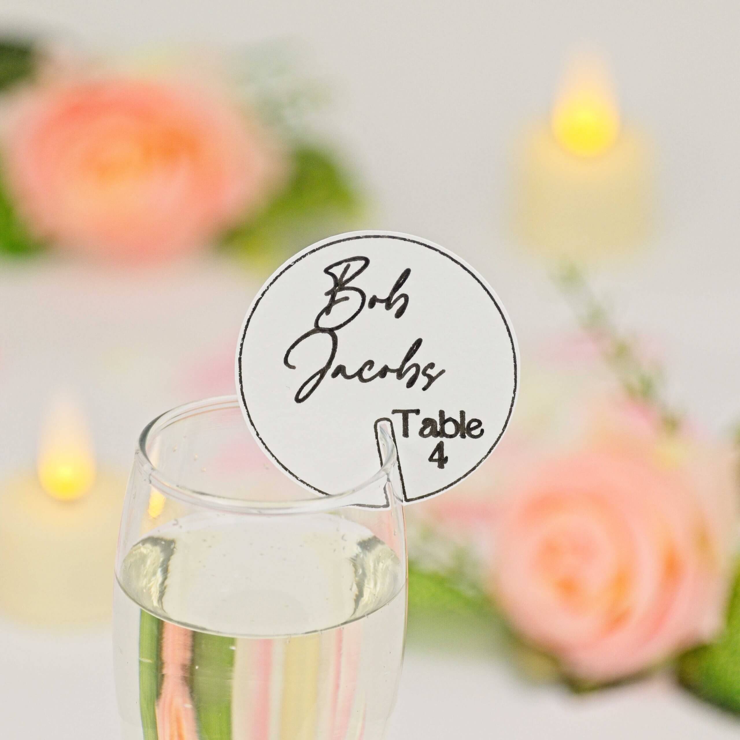 Round white drink tag with black script guest name and "Table 4", slotted on a champagne flute; out-of-focus roses and candles in background.