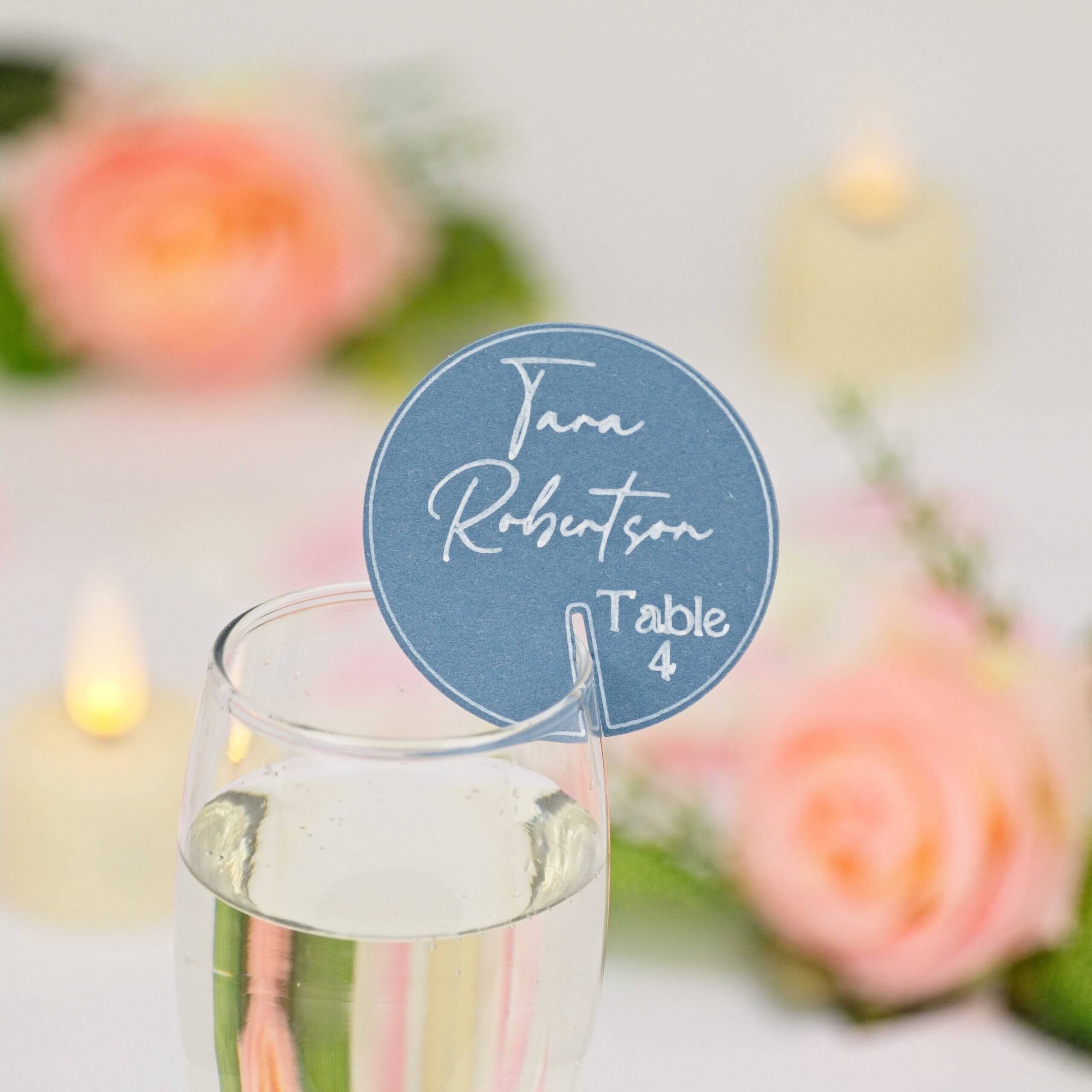 Round denim-blue drink tag with white script guest name and "Table 4", clipped to a champagne flute; blurred peach roses and candlelight behind.