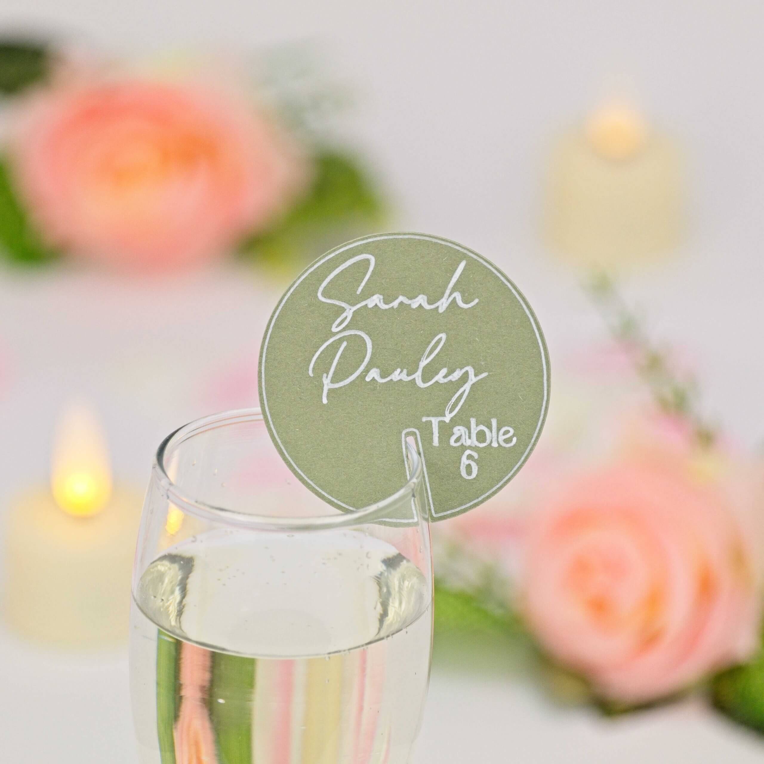 Round sage-green drink tag with white script guest name and "Table 6", slotted on a champagne flute; soft-focus rose and candle decor in background.