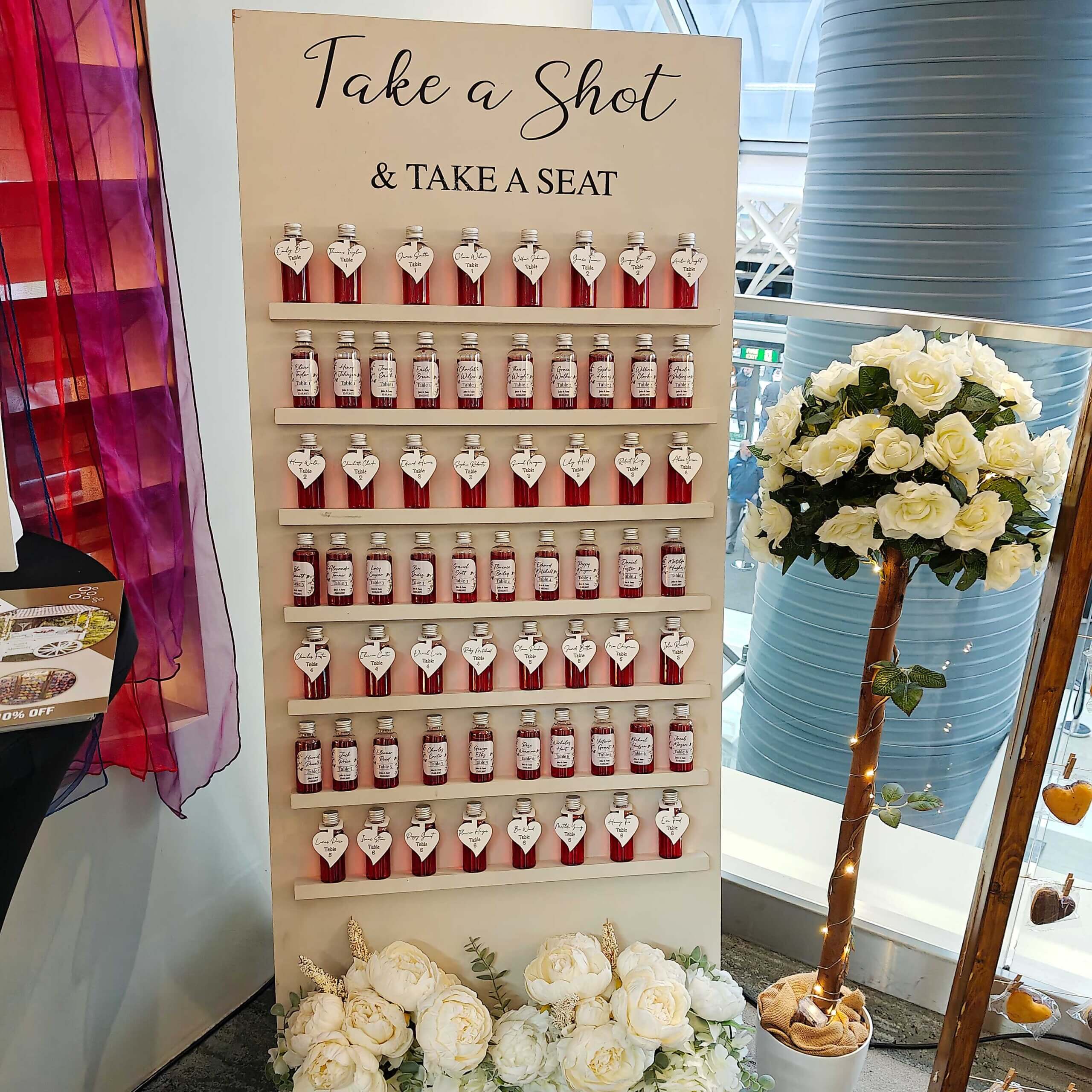 Shot wall holding 64 plastic miniature shot bottles, filled with a red liquid and adorned with square labels and/or heart shaped tags.