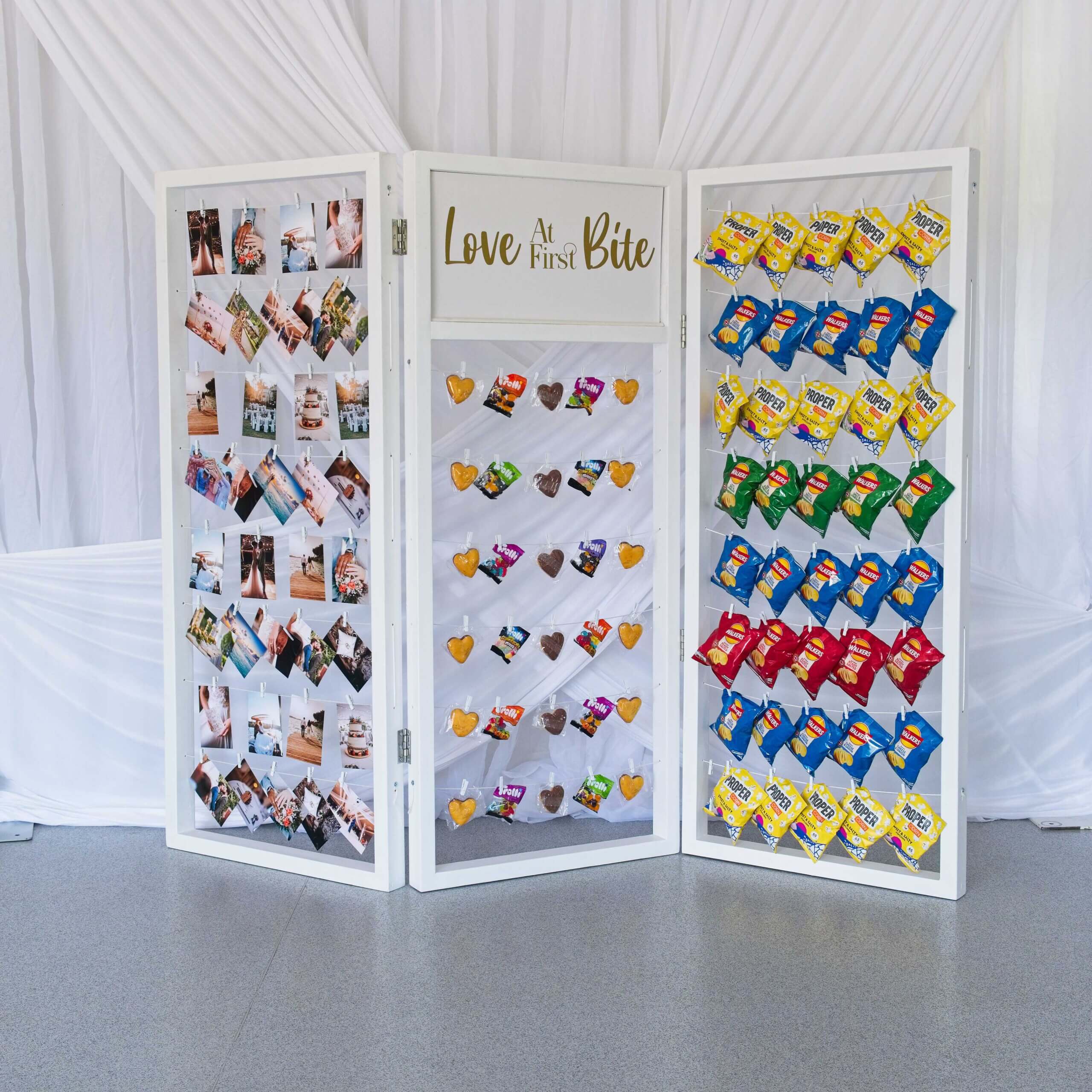 White crisp wall with crisps hanging on string lines with pegs. Photos are also hung up as well as some cakes and sweets. A sign in the centre says love at first bite.