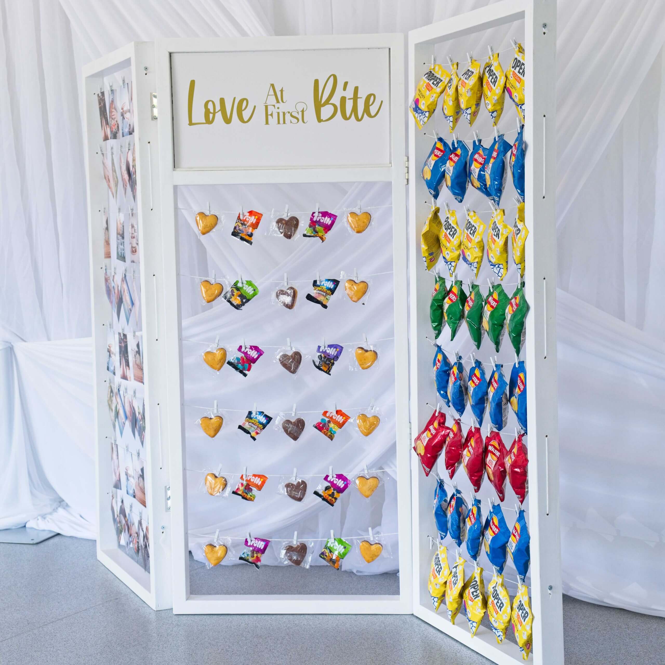 White crisp wall with crisps hanging on string lines with pegs. Photos are also hung up as well as some cakes and sweets. A sign in the centre says love at first bite.