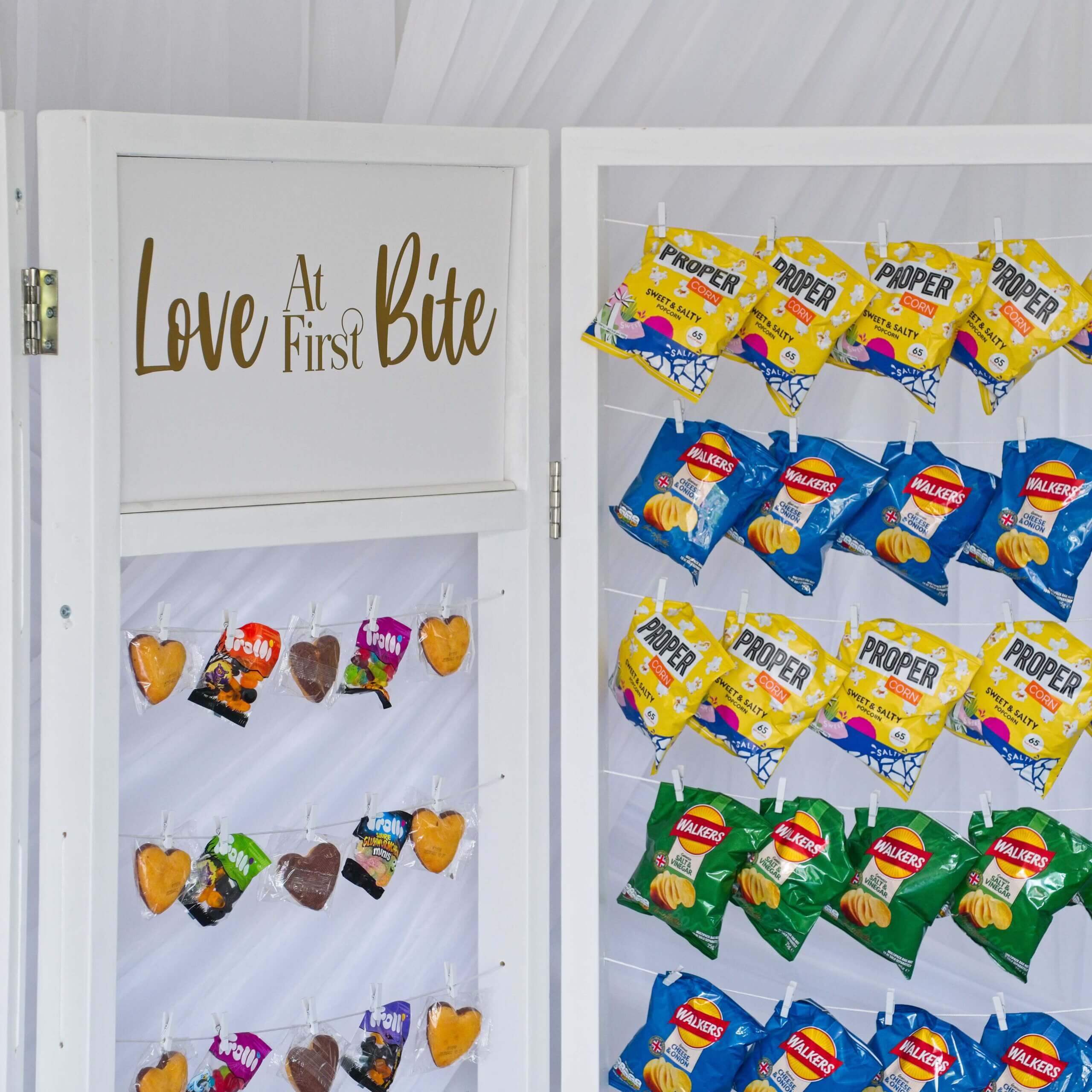 White crisp wall with crisps hanging on string lines with pegs as well as some cakes and sweets. A sign in the centre says love at first bite.