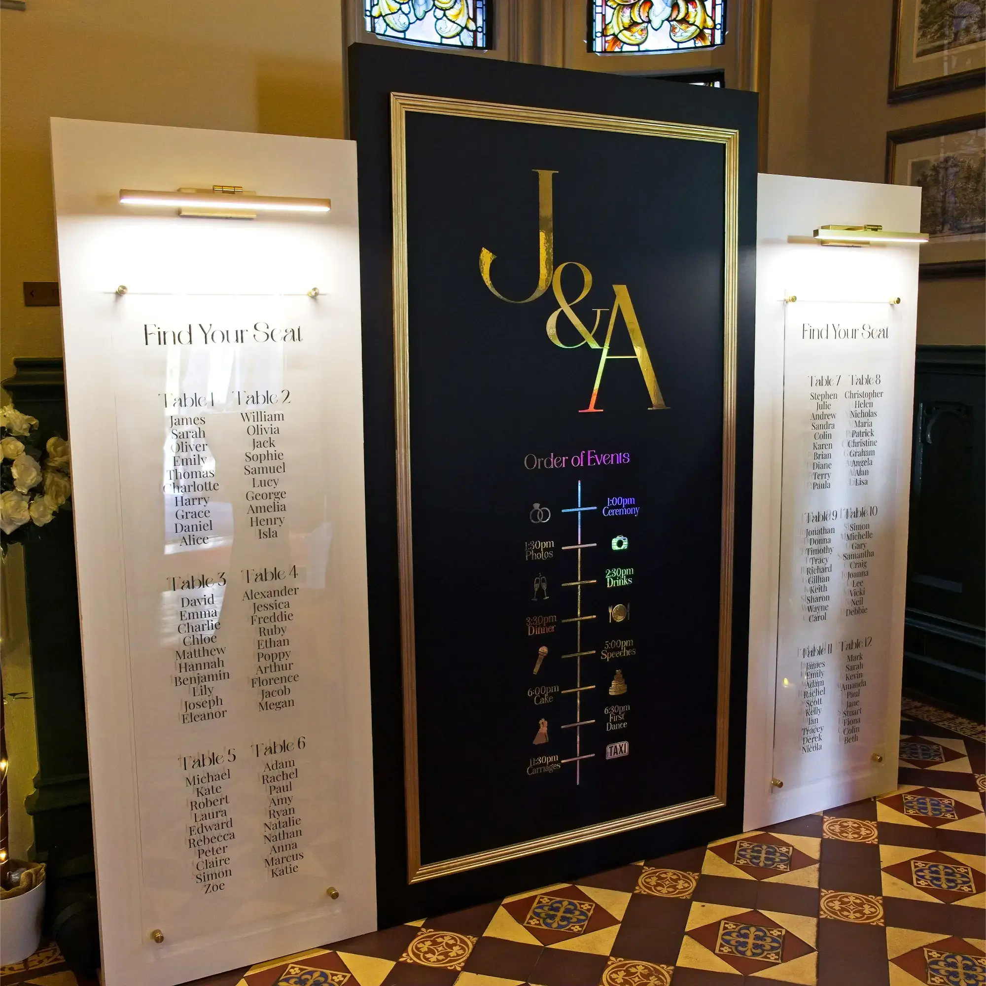 Luxury Wedding Welcome Sign & Seating Plan Display. Three panels with a central black panel with gold accents. Couple's initials in gold writing with the order of events below. Two white side panels hold top down lights and clear acrylic with a seating plan in black text.