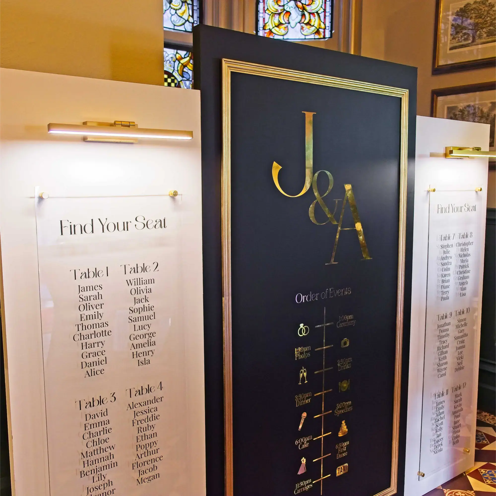Luxury Wedding Welcome Sign & Seating Plan Display. Three panels with a central black panel with gold accents. Couple's initials in gold writing with the order of events below. Two white side panels hold top down lights and clear acrylic with a seating plan in black text.