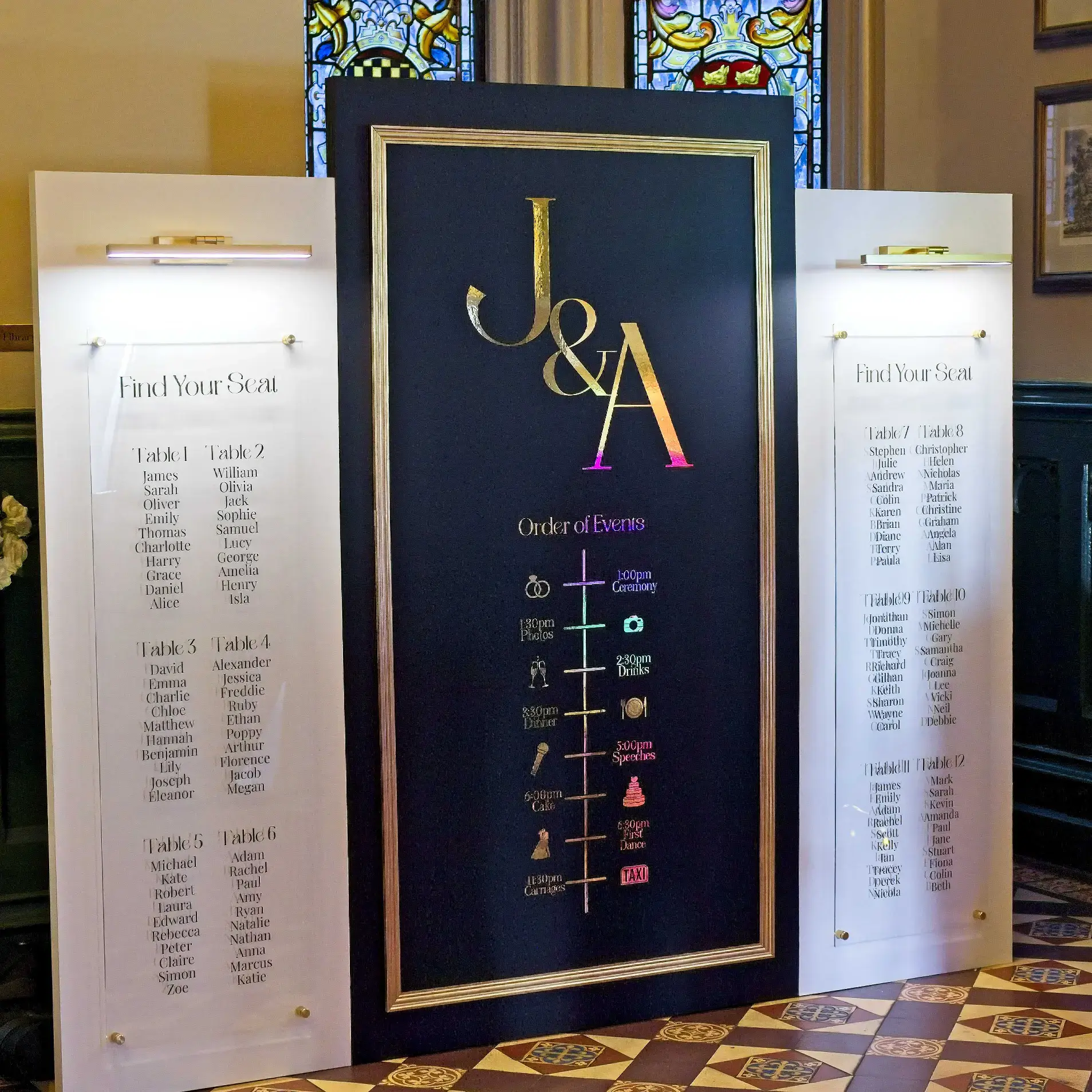 Luxury Wedding Welcome Sign & Seating Plan Display. Three panels with a central black panel with gold accents. Couple's initials in gold writing with the order of events below. Two white side panels hold top down lights and clear acrylic with a seating plan in black text.